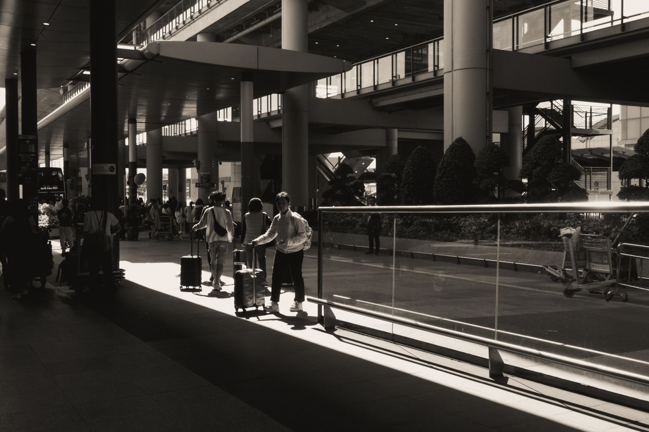 ours-journey Silhouettes of travelers with luggage at a sunlit modern airport terminal in black and white.